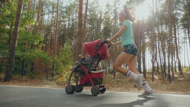 Running woman with baby stroller enjoying a sunny summer day in a pine forest. Jogging or power walking supermom, active family with baby jogger. Slow-motion 4k video