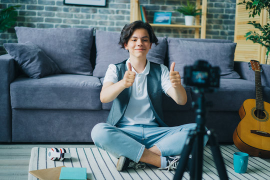 Creative boy showing thumbs-up sitting on floor in front of camera recording video for internet vlog. Modern lifestyle, happiness and people concept.