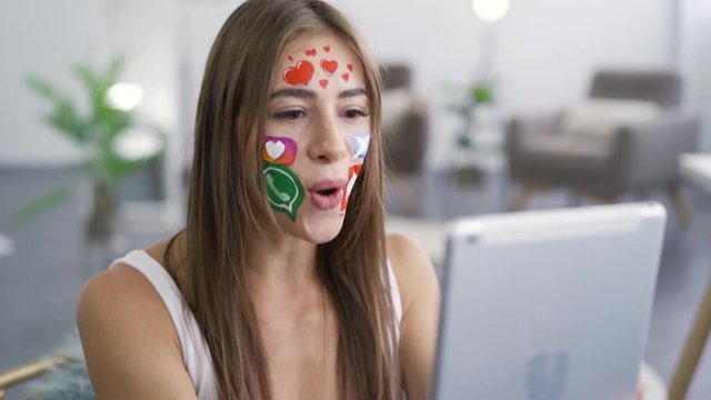 Portrait of young attractive woman with painted social media icons on her face talking with her friend using camera on her tablet. Addiction to gadgets