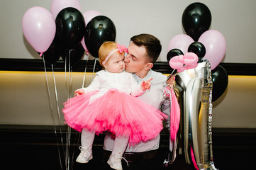 Father holding and kissing daughter in a skirt with pink tulle against a background of black and pink balloons. Man and little baby girl princess. Concept of birthday celebration is 1 year. - Powered by Adobe