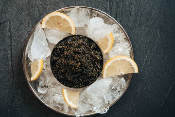 Sturgeon black caviar in can in the plate with ice and lemon on black table
