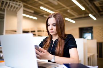 Serious business person reading text message on mobile phone during work on laptop computer. Female entrepreneur using messenger on cellphone waiting video conference on notebook. Woman using apps