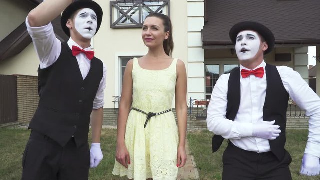Two funny male mimes with white faces in white and black clothes doing performance with young girl in front of large house. Actors making show outdoors
