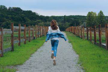 Young girl wearing a blue jeans and jeans jacket running outdoor. Girl running along the road in the stable. People, freedom, happiness concept. Full length of a happy young girl running   outdoors.