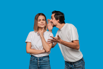 Bearded man with mustaches in white t-shirt tells secret to a young blond girl in white t-shirts and blue jeans standing isolated over blue background. Concept of secret telling.