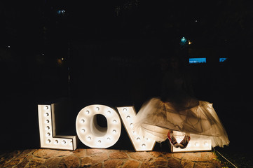 Bride seated on the word love in the evening. Word love consisting of the letters highlighted with bulbs on stone at night. Love word letters in light. The word love made with lights in a dark night.
