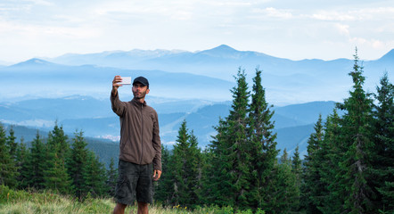 hiker man in cap making selfie in front of mountain range covered with blue fog in carpathian mountains