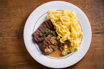Grilled beef steaks and noodles in creamy mushroom sauce, garnished with thyme on white plate