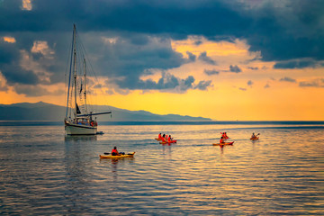 Yacht in the mediterranean sea at sunrise, Cagliari, Italy. People sail on kayaks on the open sea under the sun. The sun illuminates the island with beautiful golden colors. - obrazy, fototapety, plakaty