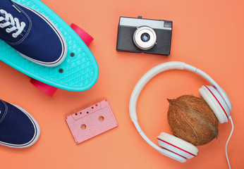 Hipster fashion look. Skateboard, sneakers, retro camera, coconut with headphones on coral color background. Top view. Flat lay
