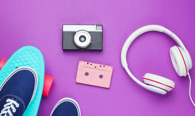 Hipster fashion look. Skateboard, sneakers, audio cassette, retro camera, headphones on purple color background. 80s. Top view. Flat lay