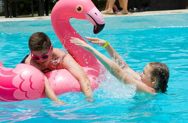 Teenage girl and boy playing with inflatable pink flamingo in luxury hotel swimming pool. Summer vacation concept