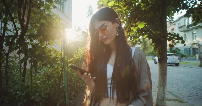 Portrait of annoyed young brunette woman using mobile phone social media reading bad news standing on street at sunset.