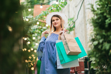 Happy shopping concept. Cheerful young attractive woman in a hat and dress holds shopping bags in her hands and smiles while walking along the street