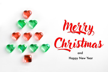 Christmas tree made of red and green decorative glass hearts laying on white background. With text Merry Christmas and Happy New Year. Holiday greeting cards, invitations, blog posts, banners design.