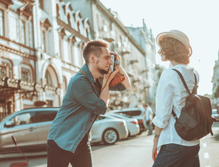 Obraz na płótnie Canvas Young cheerful couple of hipsters are photographed on a retro camera while walking around the city. Male photographer photographs a stylishly dressed young woman