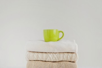 A green cup with a hot drink (coffee, tea, cocoa, cappuccino) on a stack of neatly folded knitted sweaters on a beige background. The concept of comfort, warmth, coziness, autumn-winter season.