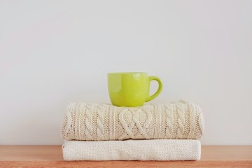 A green cup with a hot drink (coffee, tea, cocoa, cappuccino) on a stack of neatly folded knitted sweaters on a beige background. The concept of comfort, warmth, coziness, autumn-winter season.