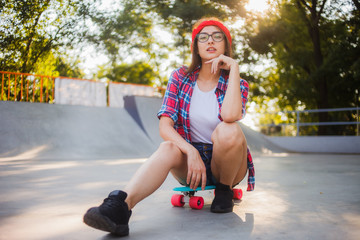Young stylish woman dressed in youth clothes sits on a skateboard at a skatepark. Summertime, sunny bright day