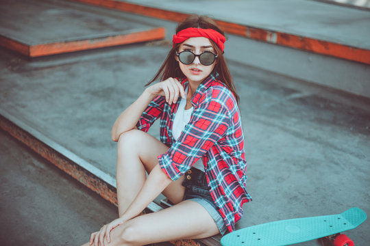 Portrait of stylish skater woman in skatepark. Attractive girl in red plaid shirt, mirrored sunglasses, shorts rests with a skate. Summer lifestyle image of trendy pretty young girl