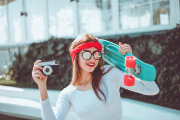 Retro style. 80s. Portrait of stylish dressed young woman with skateboard and film camera outdoors...