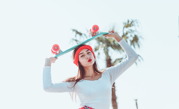 Portrait of stylish dressed young woman with skateboard outdoors on bright sunny day. Summer lifestyle image of trendy pretty young girl.