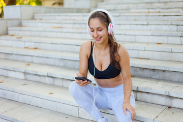 Concept of rest after training. Young happy sport woman in sportswear use smartphone and listens to music in headphones while sitting on stairs on bright sunny day