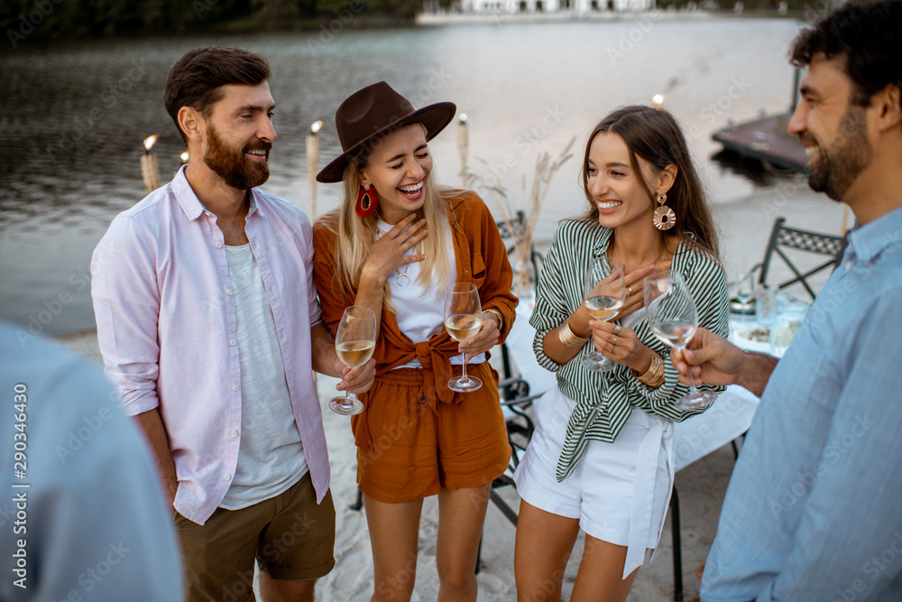 Wall mural Young friends having fun, hanging out together with wine glasses during a festive dinner on the beach at dusk