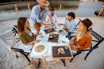 Group of friends having a festive dinner at the beautifully decorated table with delicious meals on the beach at dusk