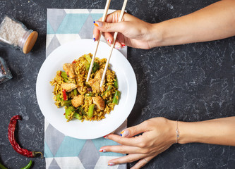 Rice with chicken meat and vegetables in a plate on stone table. Female hands hold sticks