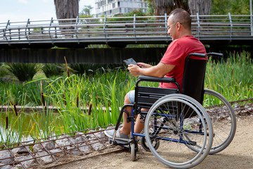 disabled man in wheelchair having fun while resting using a tablet computer at park, concept of technological and occupational integration of people with disabilities and reduced mobility problems