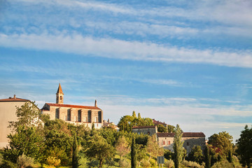 View of  the old town of Barjac, southern France. Green trees, buildings and stone church with a bell tower on a hill at sunset light