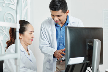 Fototapeta premium Indian male doctor showing something on tablet pc to his Asian colleague while she sitting at the table at office