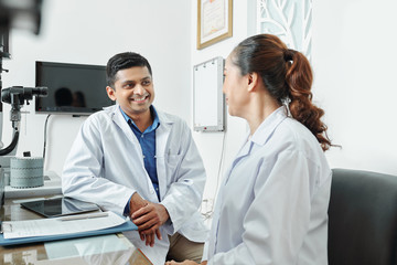 Indian mature man in white coat sitting at the table smiling and talking to the female doctor at office