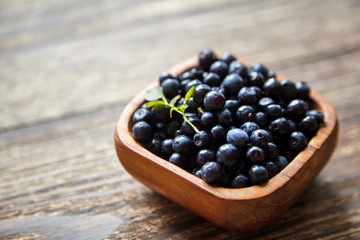 Bilberries with green leaves inside a bowl on brown wooden background. Fresh dark wild berries on table