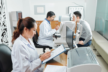 Fototapeta premium Asian nurse in white coat sitting at the table and filling the medical card while doctor examining the patient in the background at hospital
