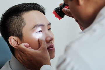 Asian young man sitting at doctor's office while optometrist examining his eyes with medical lamp
