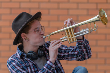 Vertical image of a boy with a hat and glasses sitting on the floor playing a yellow trumpet, with a brick wall in the background out of focus. Concept of cultural youth.