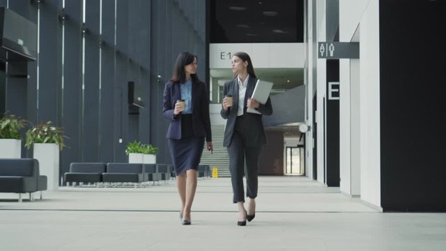 Full length dolly shot of two friendly businesswomen, young and middle aged, in formal suits walking down office hall together and talking. Colleagues holding takeaway coffee cups and laptop computer