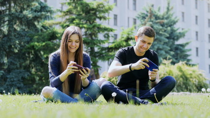 Happy friends play mobile game on smartphone, man and woman sitting on the grass in the city park.