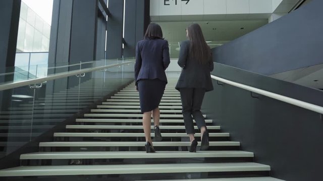 Back view follow shot of two businesswomen in formal suits and high heels walking upstairs in office building and talking. Middle aged woman holding takeaway coffee cup, young one carrying laptop