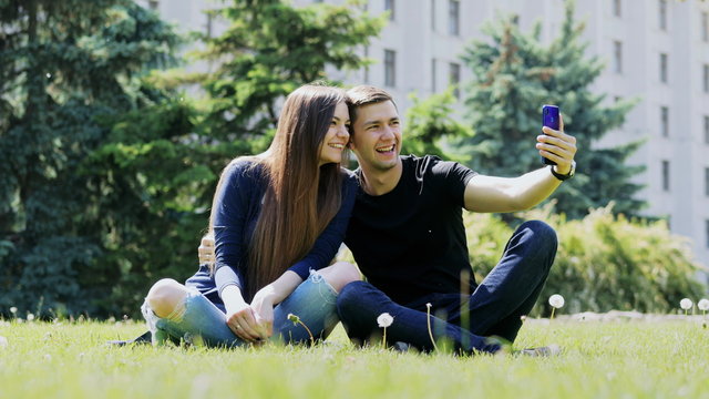 Happy couple taking selfie using smartphone. Man and woman spending time in the city park, sitting on the grass