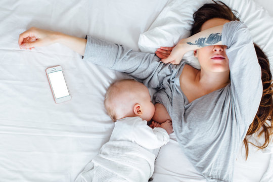 A tired mother covering her eyes as she breastfeeds her baby boy lying down on a bed. Her phone is next to her.