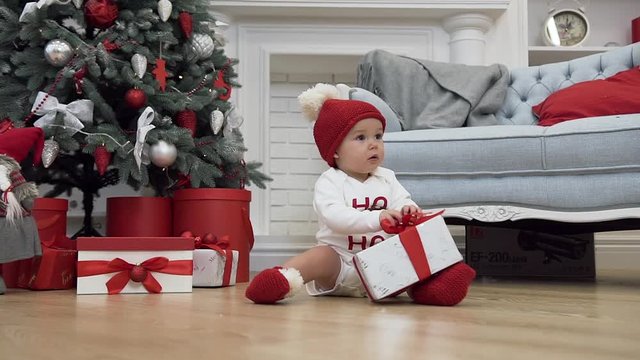 Attractive picture of smiling baby in christmas clothes sitting on the wooden floor and playing with a gift
