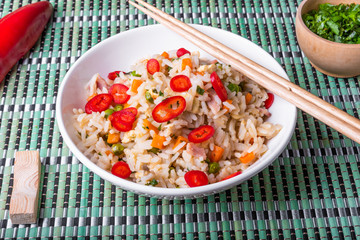 Fried rice with egg, ham and vegetables sprinkled with hot chili peppers - a traditional Asian dish of fried rice, eggs, ham, carrots and spices in a bowl on a bamboo napkin, chopsticks, close-up