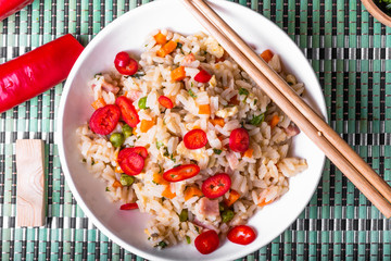 Fried rice with egg, ham and vegetables sprinkled with hot chili peppers - a traditional Asian dish of fried rice, eggs, ham, carrots and spices in a bowl on a bamboo napkin, chopsticks, close-up