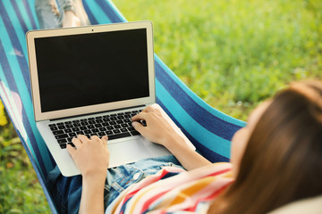 Young woman with laptop resting in comfortable hammock at green garden