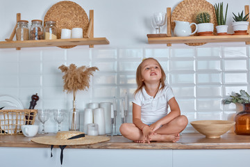 A smiling little girl sitting on the work surface of the kitchen waiting for breakfast. Cheerful and mischievous girl in the kitchen