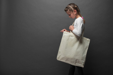 Kid girl holding white textile eco bag against gray background. Ecology or environment protection...