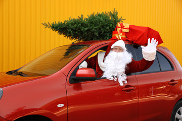 Authentic Santa Claus with fir tree and bag full of presents driving car against yellow background